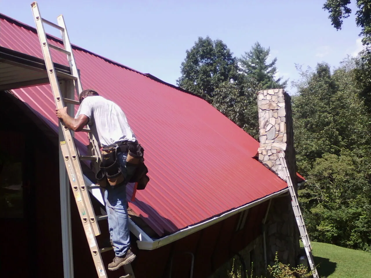 Roofer on a ladder beside a red metal roof and stone chimney — Soffit & Fascia Repair by Roofing Brothers in Roxobel